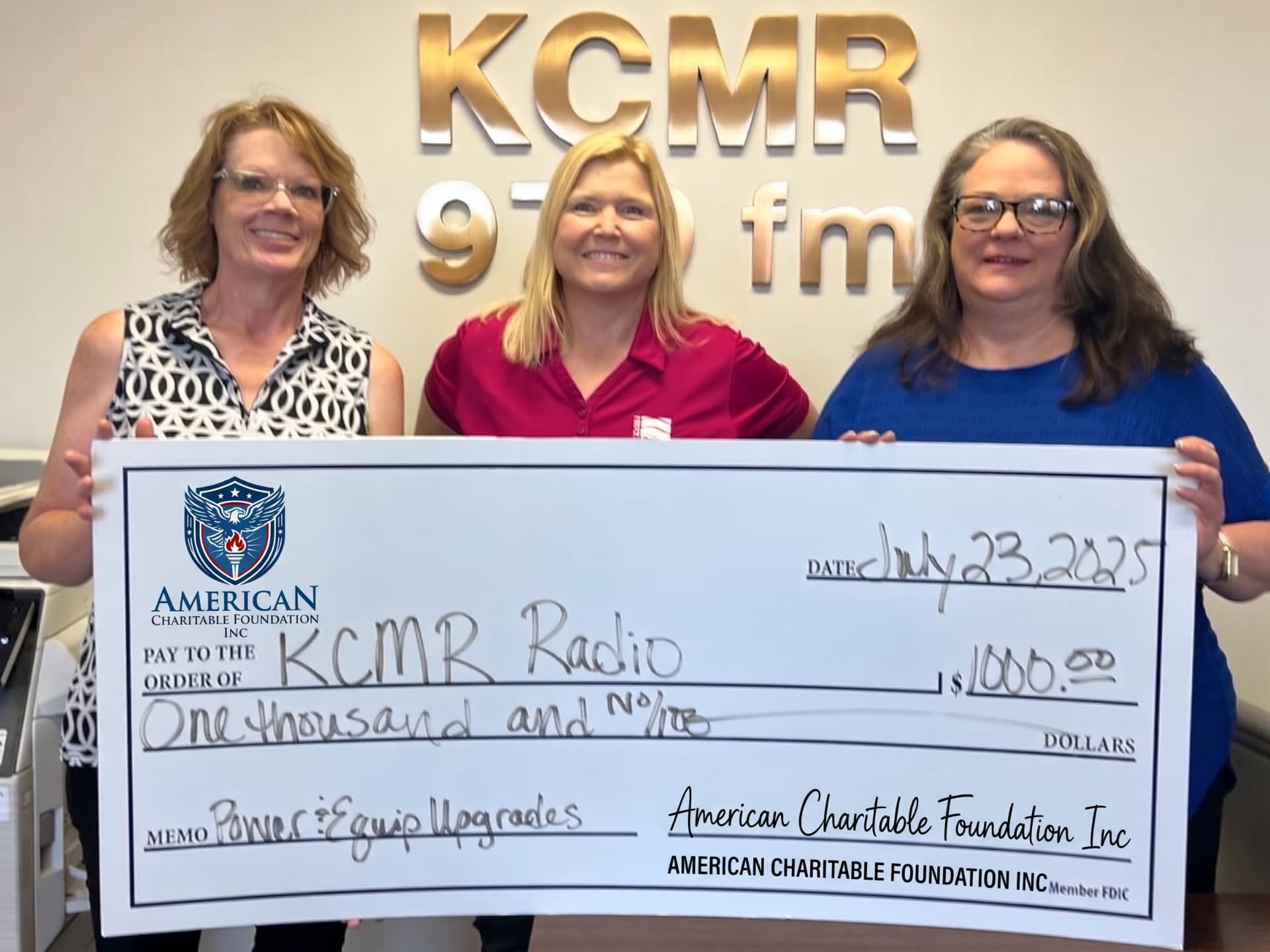 Three women presenting a large ACF grant check for KCMR Radio inside a radio station studio