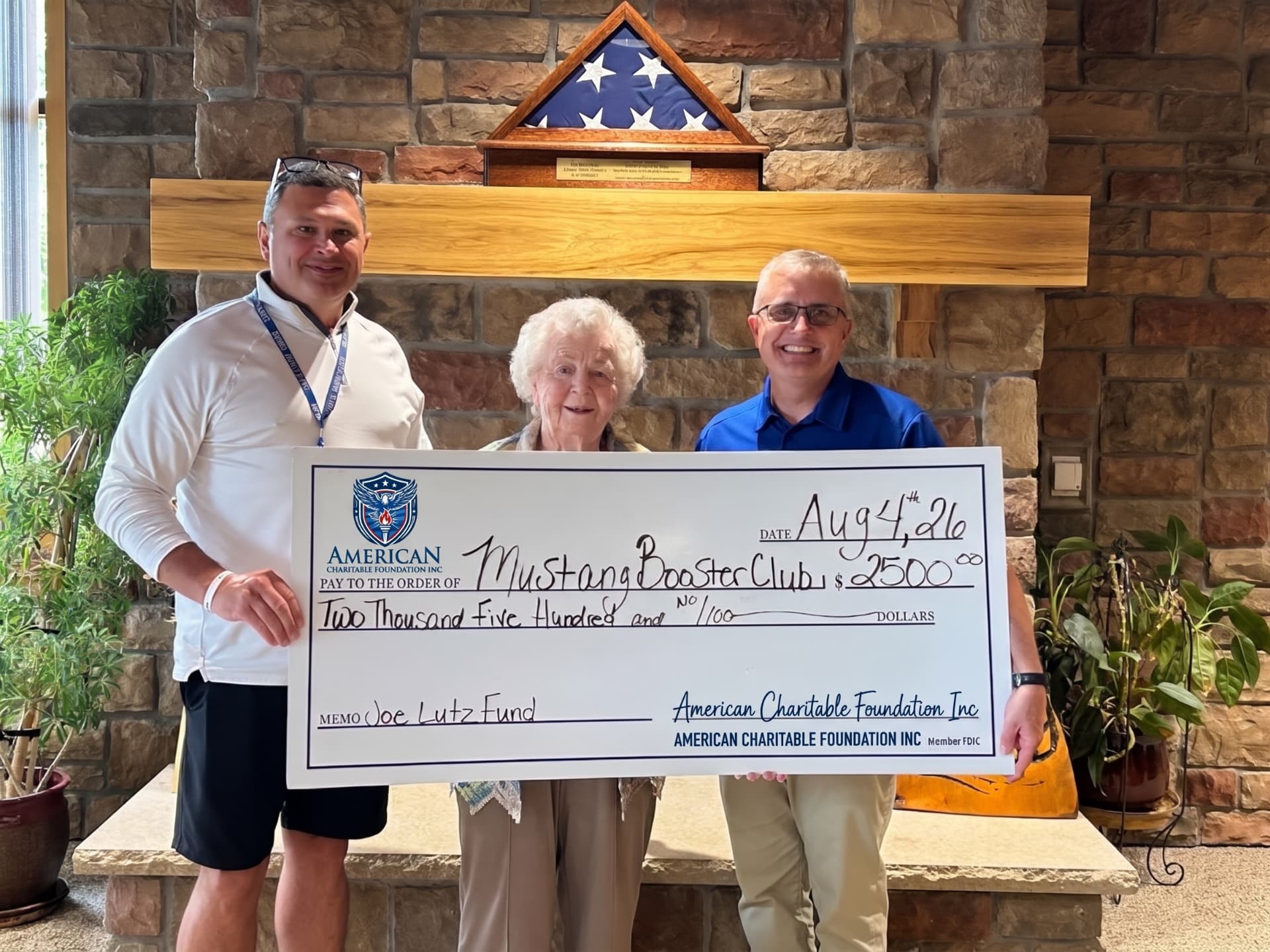 Three people presenting a large ACF grant check for the Mustang Booster Club beside a stone fireplace