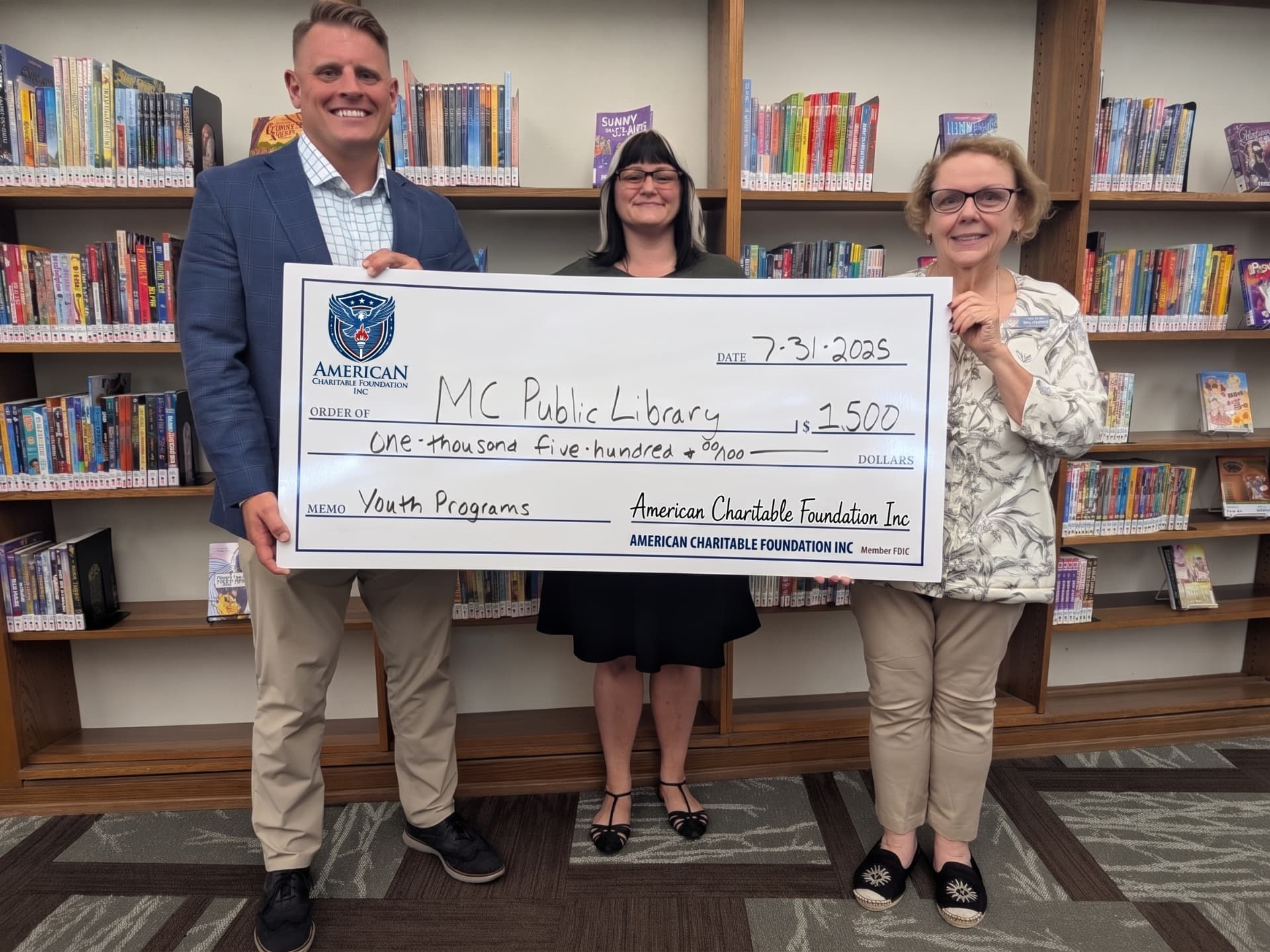 Three people presenting a large ACF grant check for MC Public Library in front of library bookshelves