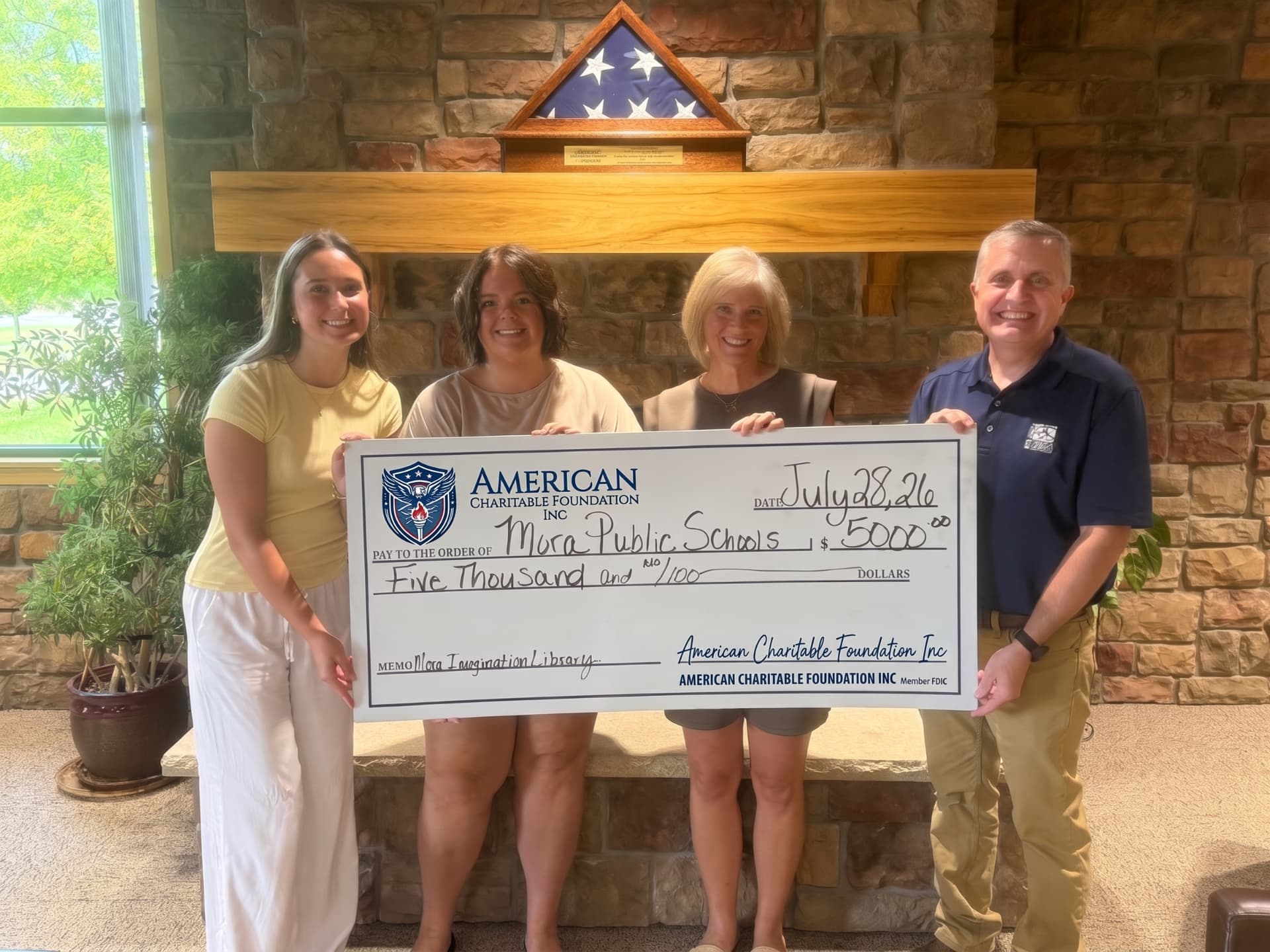 Four people presenting a large ACF grant check for Nora Public Schools beside a stone fireplace and a folded flag case